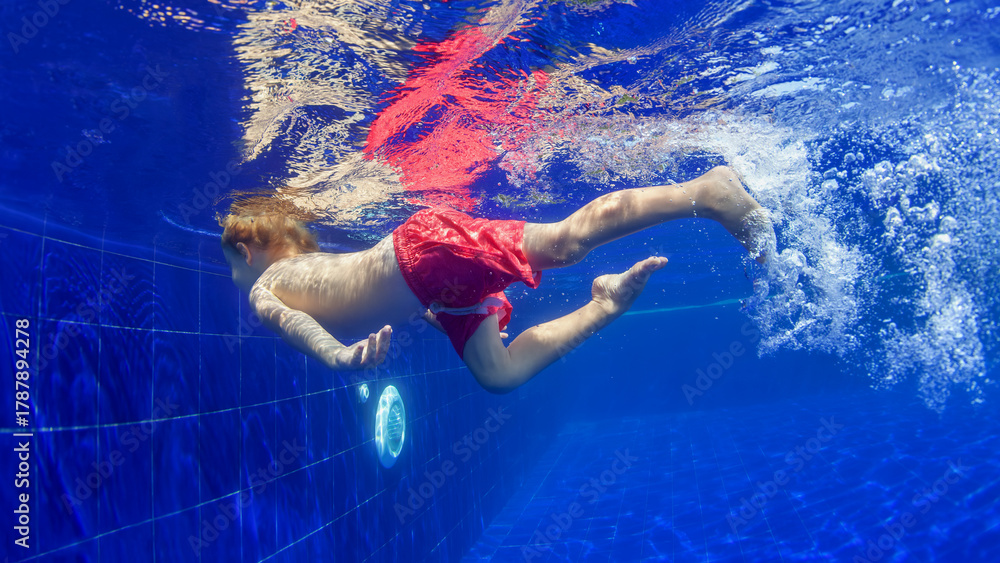 Naklejka premium Underwater shot of happy smiling young child jumping with splashes and diving with fun in blue swimming pool. Healthy family lifestyle, water sport activity, swimming lessons on holidays with kids