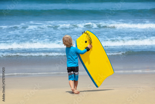 Little surfer boy with bodyboard have fun on beach, walk by white sand beach. Summer adventures, active families, parents and kids lifestyle, sports activities on school holiday with kids