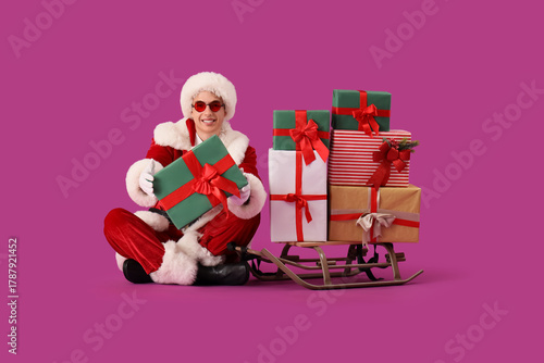 Young Santa Claus with sled and Christmas presents sitting on violet background