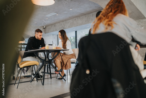 A stylish cafe scene shows a man and woman seated at a round table, discussing work over beverages. A blurred passerby adds a dynamic, candid feel to everyday business life.
