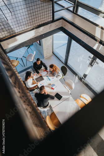 A high-angle view shows a diverse team gathered around a white table, reviewing reports and charts. Collaboration, planning, and teamwork drive a productive business meeting in a bright, open space.