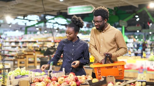 African American family couple customers buyers chooses fruits in a grocery store, market or supermarket. Man and woman makes daily purchases buy in retail shop store with basket select pick products
