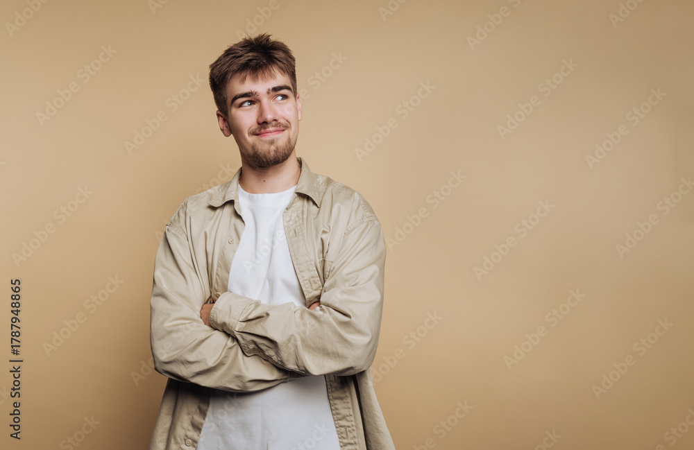 Fototapeta premium Young man with confident expression poses against neutral background while crossing arms in a relaxed manner