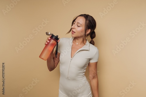 Young woman holding water bottle in light-colored athletic outfit against neutral background during workout session