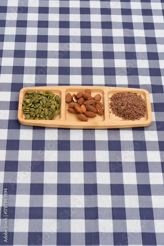 Top view of a segmented wooden tray with almonds, pumpkin seeds, and flax seeds on a navy blue and white plaid tablecloth. Vertical orientation. Focus on healthy snacks and nutrition.