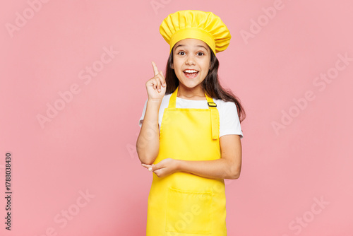 Small kid child girl housewife chef cook baker mother's helper wear yellow apron toque cap holding index finger up with great new idea isolated on pastel plain pink background. Cooking food concept.