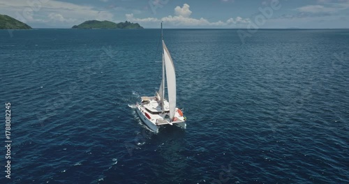 Catamaran sailing in the ocean near Kuata island, Fiji, aerial drone footage capturing the vastness of the sea and the beauty of the tropical islands in the background. Drone flight footage