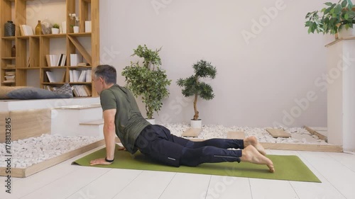 Man in sportswear practices yoga on green mat at home, performing calm stretching and strength poses including cobra