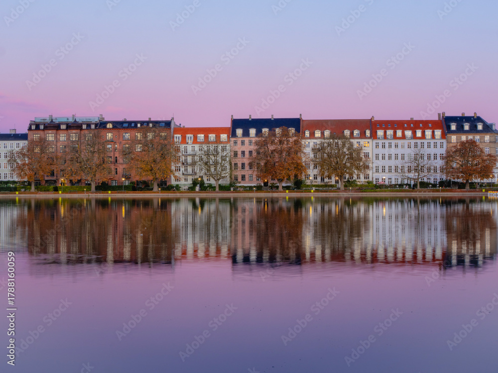 Obraz premium Colorful historic buildings reflected in calm waters at sunrise on Peblinge Sø, Copenhagen, Denmark, showcasing serene autumn cityscape in soft pastel light.