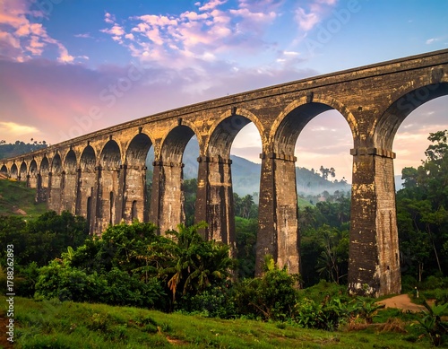 Ancient stone railway viaduct spans lush green landscape under twilight sky.