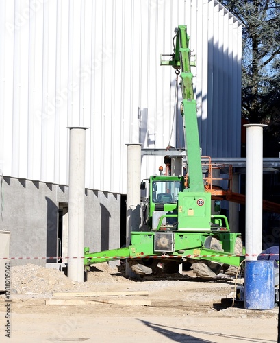 Green telescopic handler at work with a modern white facade on behind. Rear view.