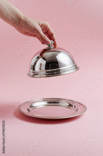 A human hand lifting a shiny silver cloche above an empty metal plate. Minimalist studio setup with a soft pink background. Concept of service, presentation, hospitality, restaurant reveal or surprise