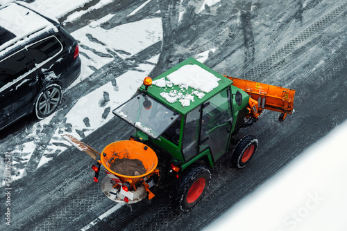 Fototapeta Naklejka Na Ścianę i Meble -  Above view small green tractor orange snow plow salt spreader clears icy street during winter in city. Fresh tire marks footprints on gray asphalt contrast with white snow and bright machine colors.