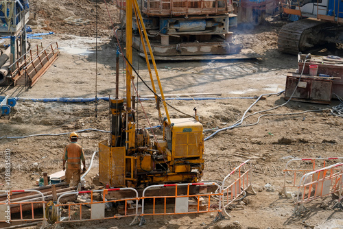 Valokuva Workers operate a yellow drilling rig on a construction site, surrounded by heavy machinery and a fenced area