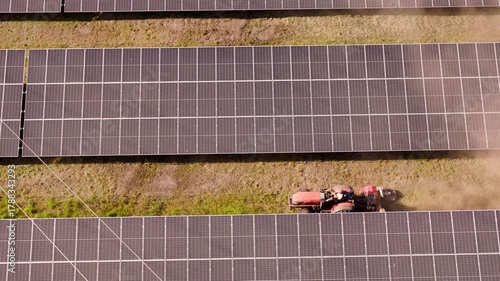 A lawn tractor cuts the grass between solar panels,drone flies over a large solar farm near a forest. Wide landscape

