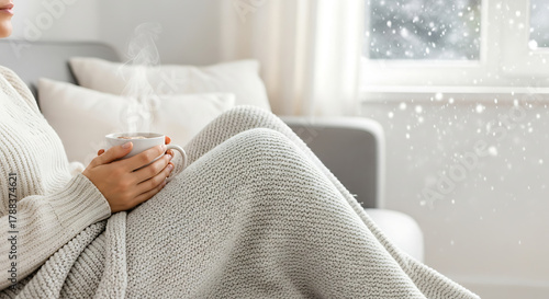 Cozy moment: a woman wrapped in a chunky knit blanket holds a steaming mug, gazing out a window as snow gently falls. Evokes hygge, comfort, and winter tranquility, ideal for wellness, lifestyle.