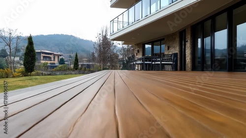 Wooden deck extends towards modern house with mountain backdrop; chairs sit by large windows