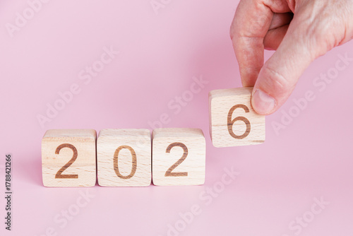 Hand is lifting wooden block with number six to replace five in sequence indicating transition from 2025 to 2026 on soft pink backdrop.