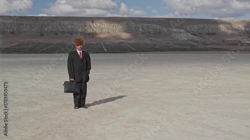 A curly-haired teenager with a sad face and a mature business suit stands on dry ground with a briefcase against the backdrop of a hill with a vineyard.
