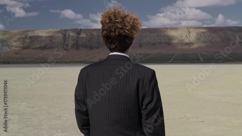 A teenager in a business suit walks against a blue sky, curls fluttering in the wind. View from behind