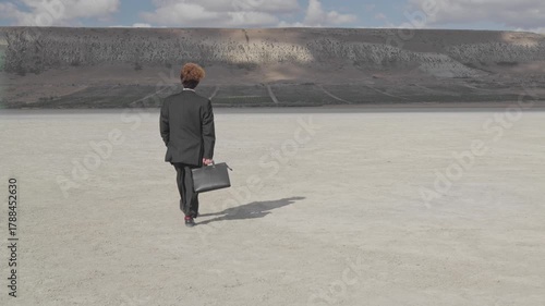 A curly-haired teenager in a business suit, carrying a briefcase, walks across dry ground on a hill with a vineyard.