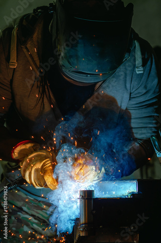 Welding shop worker in protective helmet and gloves. Blue light of welding work with bright sparks.