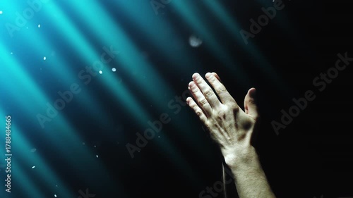 A man clasps his hands in prayer, enveloped in blue divine light and fairy dust falling from above. 4K conceptual slow motion video 60 fps.