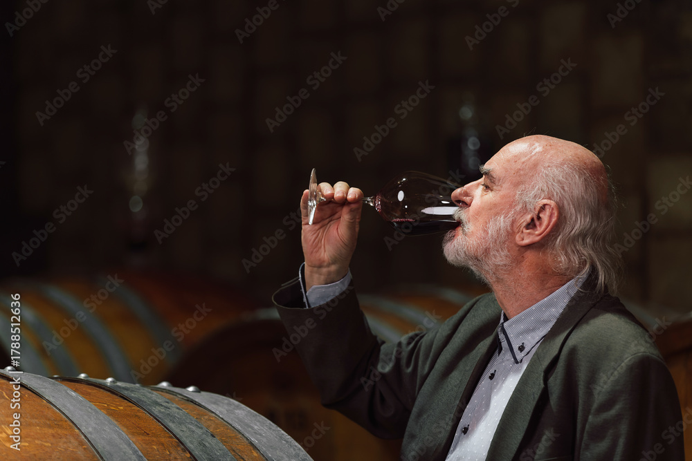 Naklejka premium A man savors a glass of red wine, surrounded by wooden barrels in a cozy winery, enjoying the rich flavors.