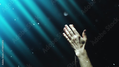 A man clasps his hands in prayer, enveloped in blue divine light and fairy dust falling from above. 4K conceptual slow motion video 60 fps.