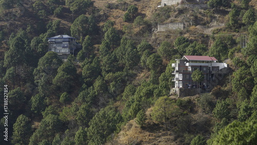 A house or structure with a light roof is perched near the crest of a steep, conical hill, primarily covered in dry, brown grass and sparse, dark green pine trees.Remote living and challenging terrain