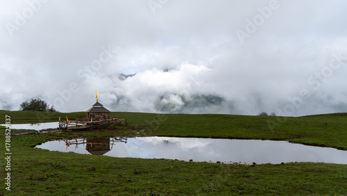 A traditional wooden temple of budha madhyamaheshwar with a distinctive tiered roof is positioned next to a still pond or lake in a lush green alpine meadow.The background is dominated by thick clouds