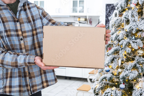 A courier with a parcel box in the kitchen with white Christmas decorations and a Christmas tree. Gift delivery for New Year and Christmas, courier service