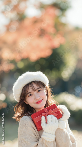 cute Japanese girl in fur beret and beige sweater smiling, hugging red present box. Outdoor winter photo in park with natural light and bokeh background.