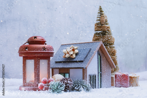 Festive winter composition with a miniature house with solar panel, glowing lantern, Christmas tree and gift boxes. Concept holiday atmosphere for Christmas, New Year and winter celebration.
