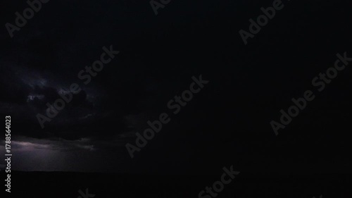Dark night sky illuminated by distant lightning flickering through storm clouds 