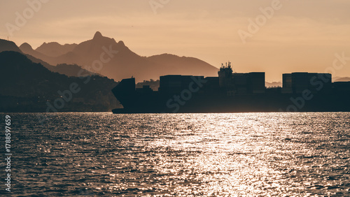 Telephoto silhouette of a large cargo ship sailing across glittering sea water at golden sunset with Rio de Janeiro mountains in the background, serene maritime scene with warm tones