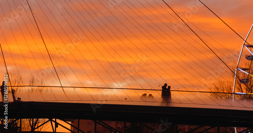 Silhouetted individuals traversing transparent pedestrian bridge during golden hour sunset, capturing urban architectural elegance against dramatic orange sky