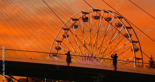 Silhouetted tourists admire illuminated Ferris wheel from pedestrian bridge during golden hour