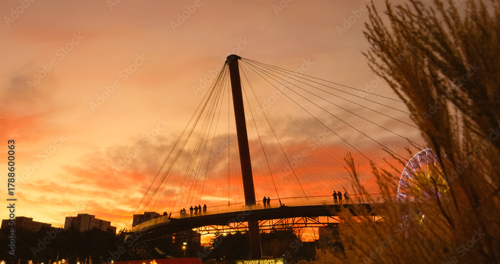 Obraz premium Pedestrians casting dark silhouettes while crossing urban bridge during golden sunset, revealing tranquil cityscape with dramatic sky and architectural elements