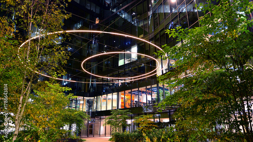 Modern office building in city center illuminated at night. Green trees near a modern office building with a glass facade. Rows of lit windows against the architectural grid of a modern facade.