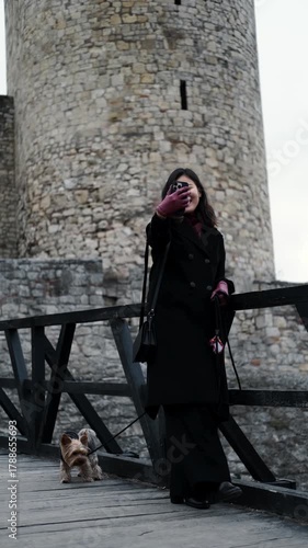 Asian woman in a black coat takes a selfie beside an old stone tower while her Yorkshire Terrier stands nearby. The concept combines travel, memory, and cultural exploration. Vertical 4k footage