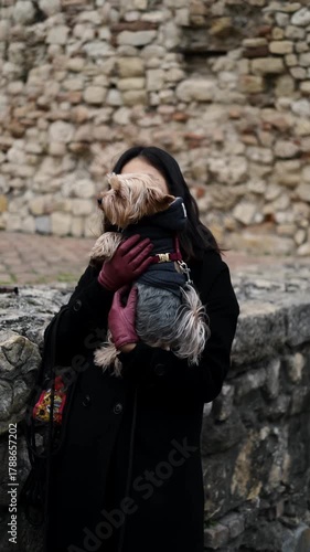 Young Asian woman in black coat pets her small Yorkshire Terrier dog near the ancient stone walls of Kalemegdan Fortress in Belgrade. Travel with pets concept. Vertical 4k footage