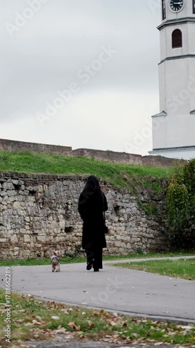 Woman walks her Yorkshire Terrier along a stone path beside an old clock tower and ancient fortress wall. The concept conveys solitude, travel, and quiet historical reflection. Vertical 4k footage