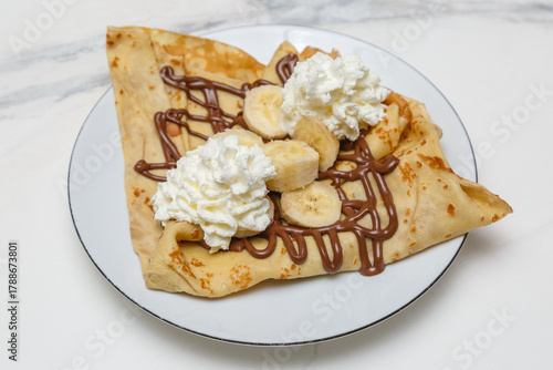 Obraz na płótnie Crêpe gourmande au Nutella, chantilly et banane, servie sur une assiette blanche sur une table en marbre