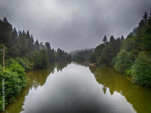 Fototapeta Naklejka Na Ścianę i Meble -  Moody, atmospheric view of a calm mountain lake surrounded by dense green forest and pine trees, with heavy fog and dark clouds obscuring the sky.Trabzon Turkey Blacksea