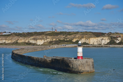 The entrance to Newhaven harbor, East Sussex, UK, and its breakwater and lighthouse. Clear blue sky and water with white cliffs in the background.