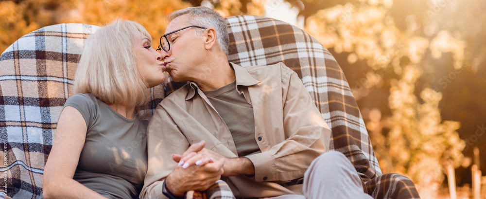 Fototapeta premium An elderly couple sits closely together in a large chair, sharing a loving kiss. The warm golden light of sunset surrounds them, highlighting their joyful moment amidst a serene outdoor scene.