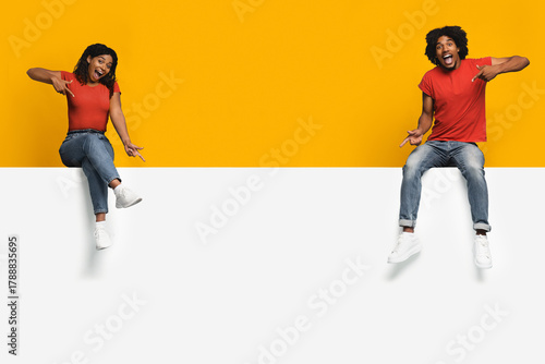 Two people sit on a ledge with bright smiles, playfully pointing at each other. They are dressed casually, with one wearing a red shirt and the other in a similar style.