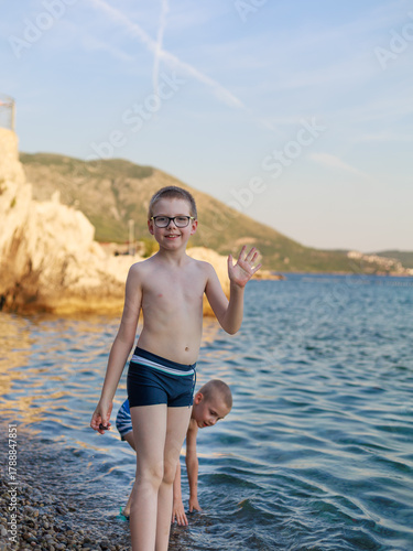 Two boys in swimwear on rocky beach at sunset, one waving and smiling to camera. Concept of summer vacation, childhood joy, travel freedom.