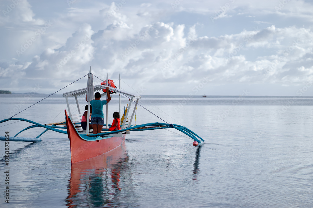 Obraz premium A family enjoys a peaceful day on a colorful red and blue outrigger boat, gliding over tranquil water as white clouds drift above, capturing togetherness and adventure at sea.